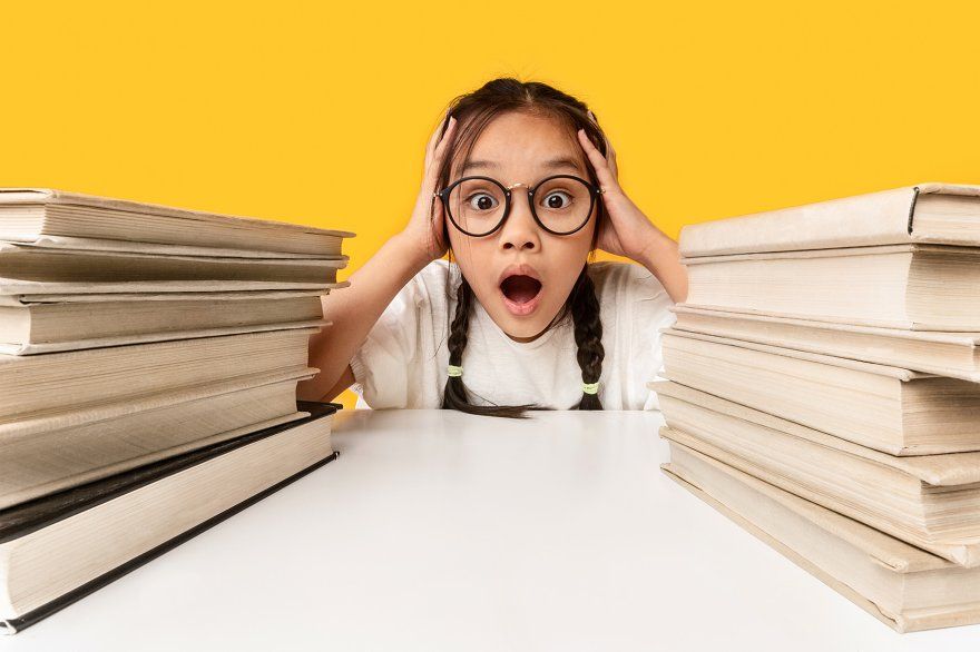 Здивована школярка між стопками книжок — перевантаження та стандартизоване тестування